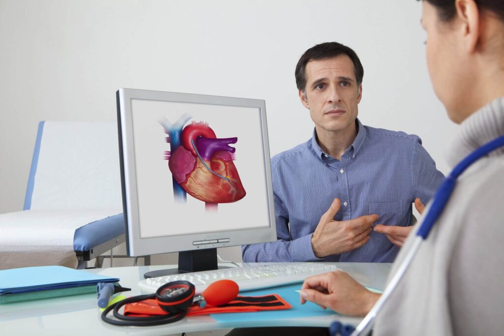 mężczyzna skarżący się na ból w klatce piersiowej, lekarz kardiolog za biurkiem spogląda na wizualizację serca znajdującą się na monitorze